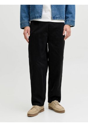 Férfi laza fekete nadrágban, bézs fűzős cipőben, fehér ingben és kék farmer dzsekiben, egyszínű világos háttér előtt áll.