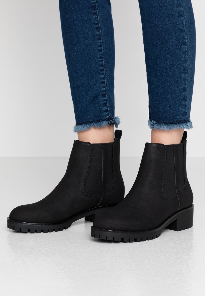 Bottes noires en cuir montant jusqu'à la cheville avec semelles épaisses, portées avec un jean skinny bleu foncé à ourlet effiloché, sur un fond clair uni.