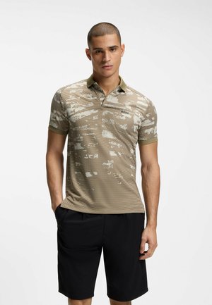 Mand med kort hår iført beige polo skjorte med mønster og sorte shorts, stående med den ene hånd i lommen mod hvid baggrund.