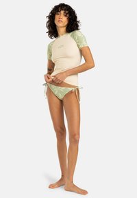 Rash guard de manga corta en beige con detalles en verde y hojas, combinado con una parte de bikini a juego que presenta lazos laterales y un patrón floral.