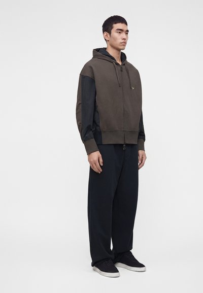 Jeune homme debout portant un sweat à capuche zippé marron foncé et noir, un pantalon noir ample et des baskets noires avec des semelles blanches sur un fond uni.