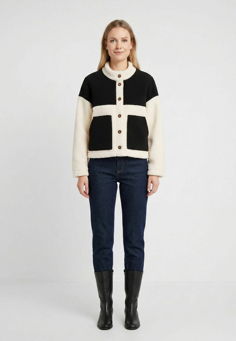 Femme portant une veste polaire boutonnée noire et blanche, un jean foncé et des bottes noires montantes, debout devant un fond uni.