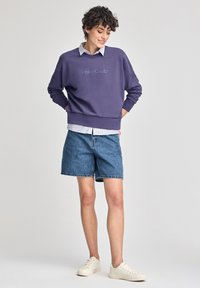 Lila sweatshirt med texten "Polo Club", i kombination med ljusa denimshorts och krämfärgade sneakers, har en lager-på-lager-look med en skjorta under.