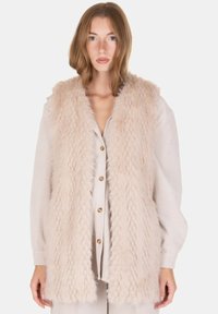 Chaleco de pelo sintético en beige claro con un diseño texturizado y de pelo largo, que presenta un cuello tipo chal y abertura frontal, llevado sobre una blusa beige abotonada.