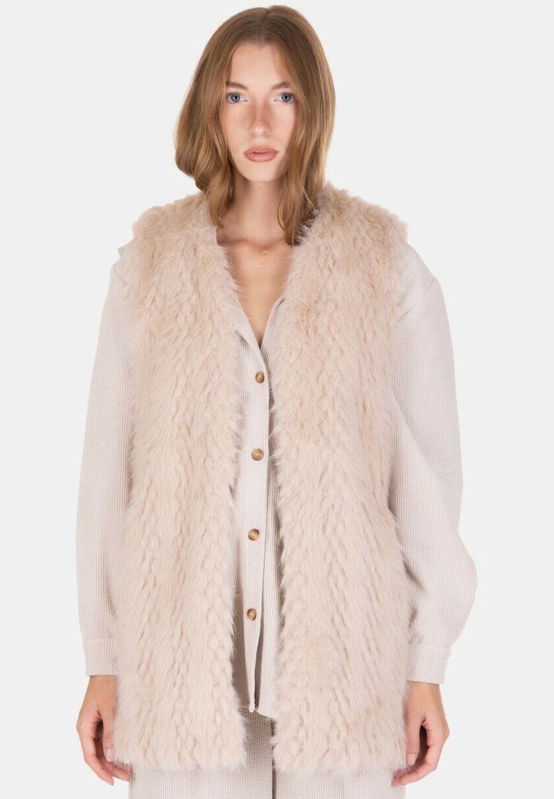 Chaleco de pelo sintético en beige claro con un diseño texturizado y de pelo largo, que presenta un cuello tipo chal y abertura frontal, llevado sobre una blusa beige abotonada.