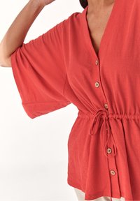 Blouse rouge à manches amples, encolure en V, fermeture par boutons, taille avec cordon de serrage, et tissu doux et texturé. Design décontracté et polyvalent.