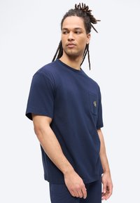 Άνδρας με dreadlocks που φοράει μπλε σκούρο κοντομάνικο T-shirt με μικρή τσέπη στο στήθος και λογότυπο, κοιτάζει προς τα αριστερά του σε απλό φόντο.