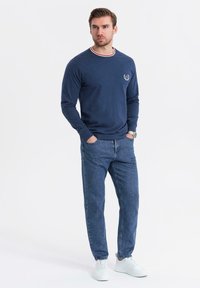 Marinefarbendes Langarmhemd mit rot-weiß gestreiftem Kragen, kombiniert mit blauen Jeans und weißen Sneakers, vor einem weißen Hintergrund stehend.