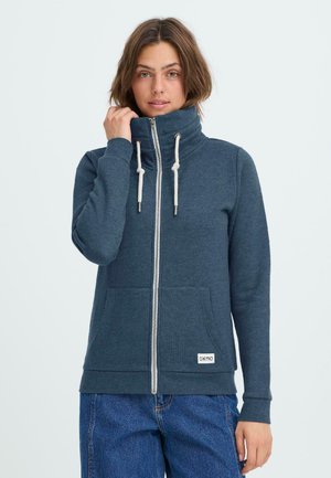 Blå zip-up hættetrøje med høj krave, snøredetaljer og forlommer. Blødt materiale med en glat tekstur og mærkeetiket på kanten.