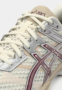 Detailní záběr na krémově zbarvenou běžeckou obuv Asics s síťovinou, béžovými tkaničkami a vínovými a stříbrnými detaily loga po stranách.