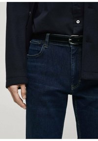 Tamni traperice s uskim krojem, koje imaju pet džepova, suptilan šav i crni remen sa srebrnom kopčom.