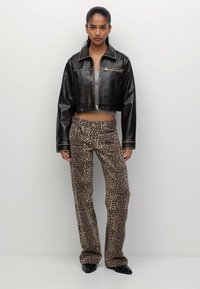 Femeie în picioare, purtând o jachetă din piele neagră scurtă, un top gri scurt, pantaloni cu imprimeu leopard și pantofi negri cu vârf ascuțit, pe un fundal simplu.