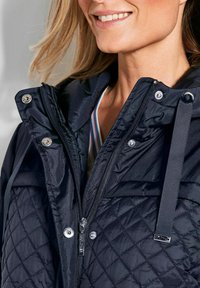 Schwarze Steppjacke mit glattem oberen Bereich, silbernen Beschlägen, einem Reißverschluss und einer Kapuze mit Kordelzug.