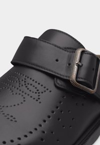 Κοντινό πλάνο μαύρου δερμάτινου κλόγ με διάτρητο σχέδιο και μεταλλικό λουράκι με πόρπη στην κορυφή.