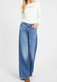 Femme portant un jean bleu à jambes larges, une chemise blanche à manches longues avec le texte « Guess », et des bottes marron à talons, debout avec une main dans la poche.