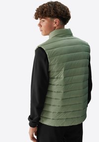 Hellgrüne Steppweste mit horizontaler Steppung, hohem Kragen und ohne Ärmel, über einem schwarzen Langarmshirt getragen. Glatte Textur.