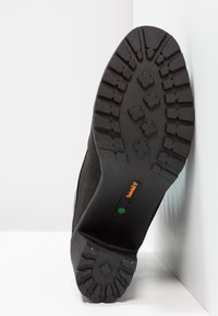 Černá podešev boty s hlubokým vzorkem, logo Timberland v oranžové barvě, zelená hexagonální insignie, umístěná vzpřímeně na bílém rohu pozadí.