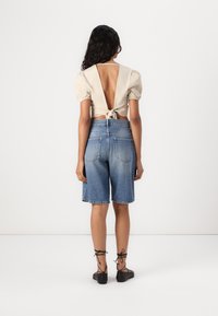 Beige puff-sleeve toppi, jossa on syvä leikkaukset selässä, sidottuna vyötäröltä, yhdistettynä keskisinisiin denimpituisiin shorts-haalareihin ja mustiin nauhakengiin.