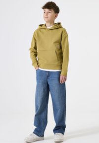 Un adolescente de pie con las manos en los bolsillos, vistiendo una sudadera verde oliva, vaqueros azules y zapatillas blancas, frente a un fondo blanco liso.