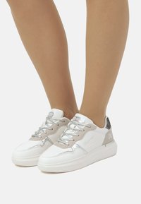 Zapatillas blancas con detalles plateados y paneles de ante beige. Presentan una suela gruesa, cordones texturizados y un logo en el lateral.