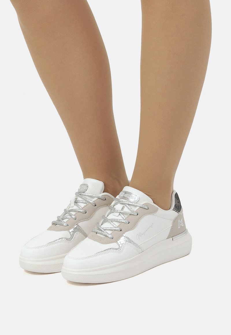 Zapatillas blancas con detalles plateados y paneles de ante beige. Presentan una suela gruesa, cordones texturizados y un logo en el lateral.