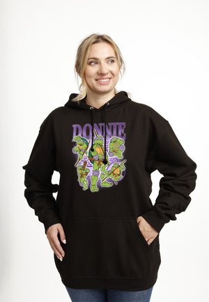 Smilende kvinde iført sort hættetrøje med farverig Teenage Mutant Ninja Turtles-karakter Donatello og teksten "DONNIE", hænderne i lommerne.