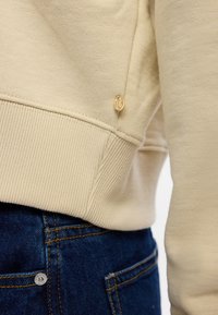 Beige mikina s ribovanými manžetami a spodním lemem, se malým zlatým logem na boku. V kombinaci s tmavými džíny.