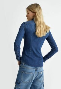Cămașă din denim de culoare albastru închis, cu mâneci lungi, guler ascuțit și manșete cu butoni. Prezintă un design ajustat pe spate, cu un tiv drept.