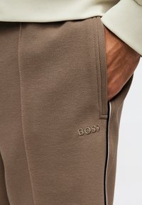 Κοντινό πλάνο ενός ατόμου που φοράει καφέ υφασμάτινο παντελόνι με πλαϊνή τσέπη και λογότυπο "BOSS", με το χέρι εν μέρει μέσα στην τσέπη και μανίκι εκρού πουλόβερ ορατό.