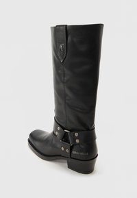 Bota de cuero negro hasta la rodilla con punta redondeada, que presenta una correa con tachuelas plateadas en el tobillo y una marca grabada en el talón.
