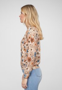 Blouse florale en tissu beige doux, ornée de fleurs bleues et oranges, à manches longues et à encolure ronde. Associée à un jean en denim clair.
