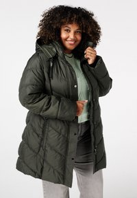 Olijfgroene pufferjack met een ritssluiting aan de voorkant, capuchon en een diagonaal geruit patroon. Heeft zwarte accenten en een relaxed fit.