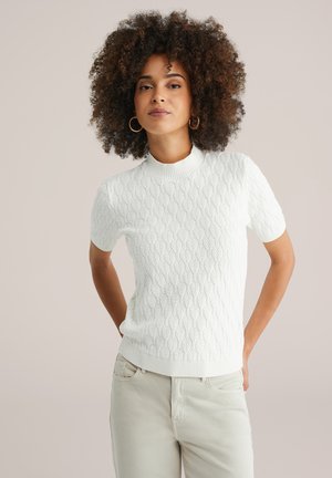Vrouw met krullend haar, draagt gouden hoopoorbellen, een witte gestructureerde trui met korte mouwen en lichtbeige broeken, tegen een effen achtergrond.