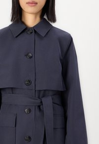 Cappotto trench blu navy con ampio colletto, sei bottoni di chiusura, cintura in vita e grandi tasche frontali. Realizzato in tessuto morbido.