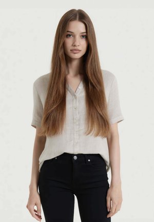 Giovane donna con lunghi capelli castani e lisci che indossa una camicia beige a maniche corte con bottoni e pantaloni neri, in piedi davanti a uno sfondo semplice.