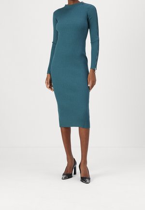 Mujer con un vestido midi de punto acanalado en color verde azulado con mangas largas y tacones altos negros, posando frente a un fondo liso y claro.