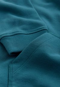 Primer plano de una sudadera azul verdoso que muestra los puños acanalados y la textura suave de la tela con detalles visibles de costuras.
