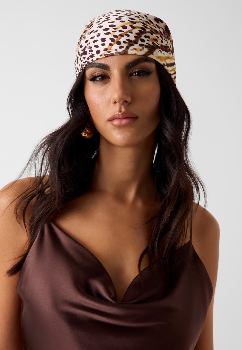 Frau mit langem dunklem Haar, die ein braunes Satinkleid und ein gemustertes Kopftuch in Weiß-, Braun- und Beigetönen trägt.