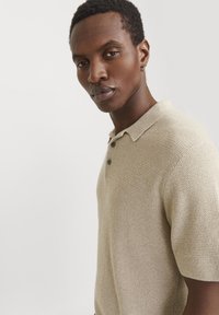 Camisa polo de manga corta en color beige con un patrón texturizado de punto, dos botones oscuros y un cuello suave con canalé.
