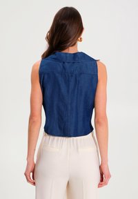 Haut sans manches en denim bleu foncé avec col, coupe ajustée et longueur courte. Texture lisse avec détails de surpiqûres le long des coutures.