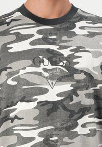 Grå kamuflasje t-skjorte med rund svart krage, med "GUESS" i svart trykk og en trekantet logo med et spørsmålstegn. Myk tekstur.
