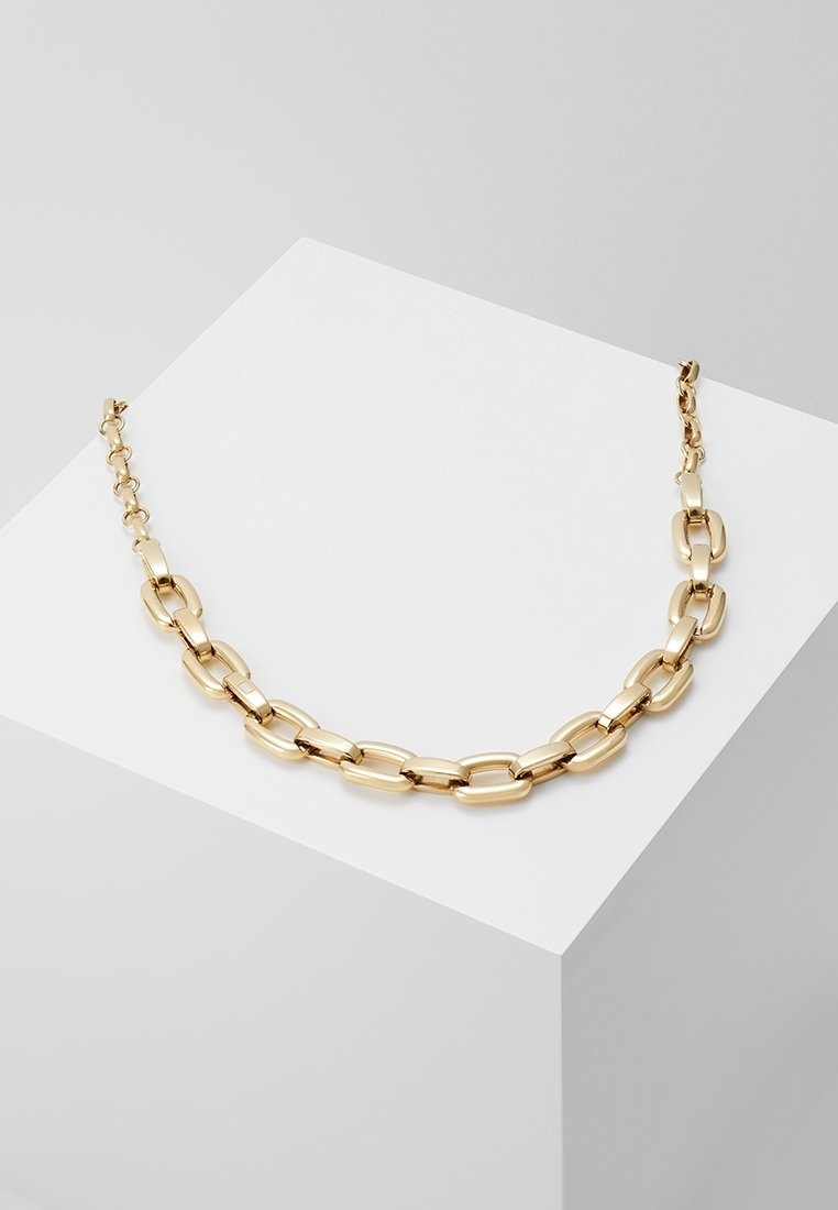 Collier chaîne en or avec de grands maillons ovales présenté sur un bloc géométrique blanc sur un fond gris.