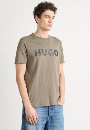 T-shirt in cotone verde oliva con scollatura rotonda, caratterizzata da un audace lettering nero "HUGO" sulla parte anteriore. Maniche corte e vestibilità rilassata.
