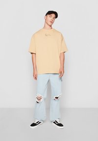 Camiseta beige de gran tamaño con mangas cortas y un logo sutil, combinada con jeans claros rasgados y zapatillas negras.