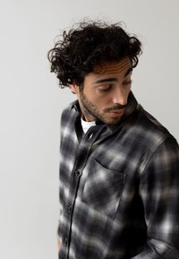 Zwart en grijs geblokt flanellen shirt met een knoopsluiting, een enkele borstzak en een gestructureerde stof. Model kijkt naar beneden tegen een neutrale achtergrond.