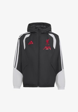 Barn med krøllet hår iført sort og hvid Liverpool FC sportjakke og bukser med røde logoer, stående mod en ensfarvet baggrund.