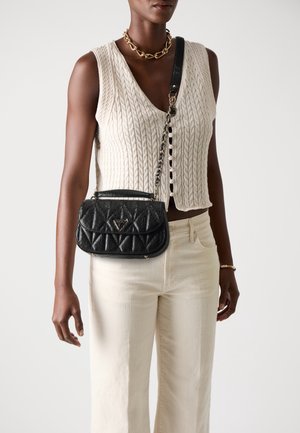 Osoba ubrana w kremowy, bezrękawnik z dzianiny warkoczowej oraz spodnie, z czarną pikowaną torbą na ramię i złotym naszyjnikiem z łańcuszkiem.