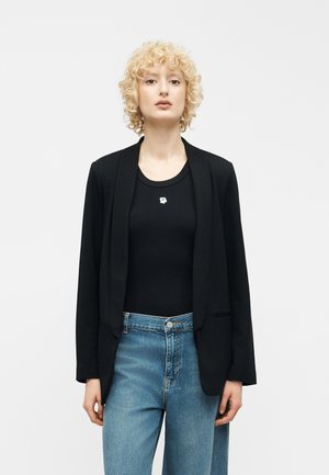 Personne blonde aux cheveux bouclés portant un blazer noir sur une chemise noire avec une petite fleur blanche, et un jean bleu, debout.