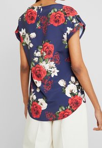Blusa floreale a maniche corte in blu navy, con rose rosse e fiori bianchi, orlo arrotondato e dettaglio con bottoni nella parte posteriore del collo.