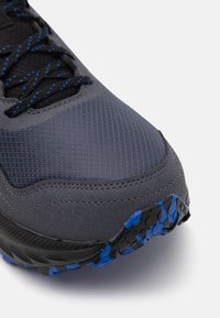 Baskets de sport grises avec un dessus texturé, présentant des accents bleus et une semelle extérieure noire robuste avec des motifs bleus, conçues pour l'adhérence.